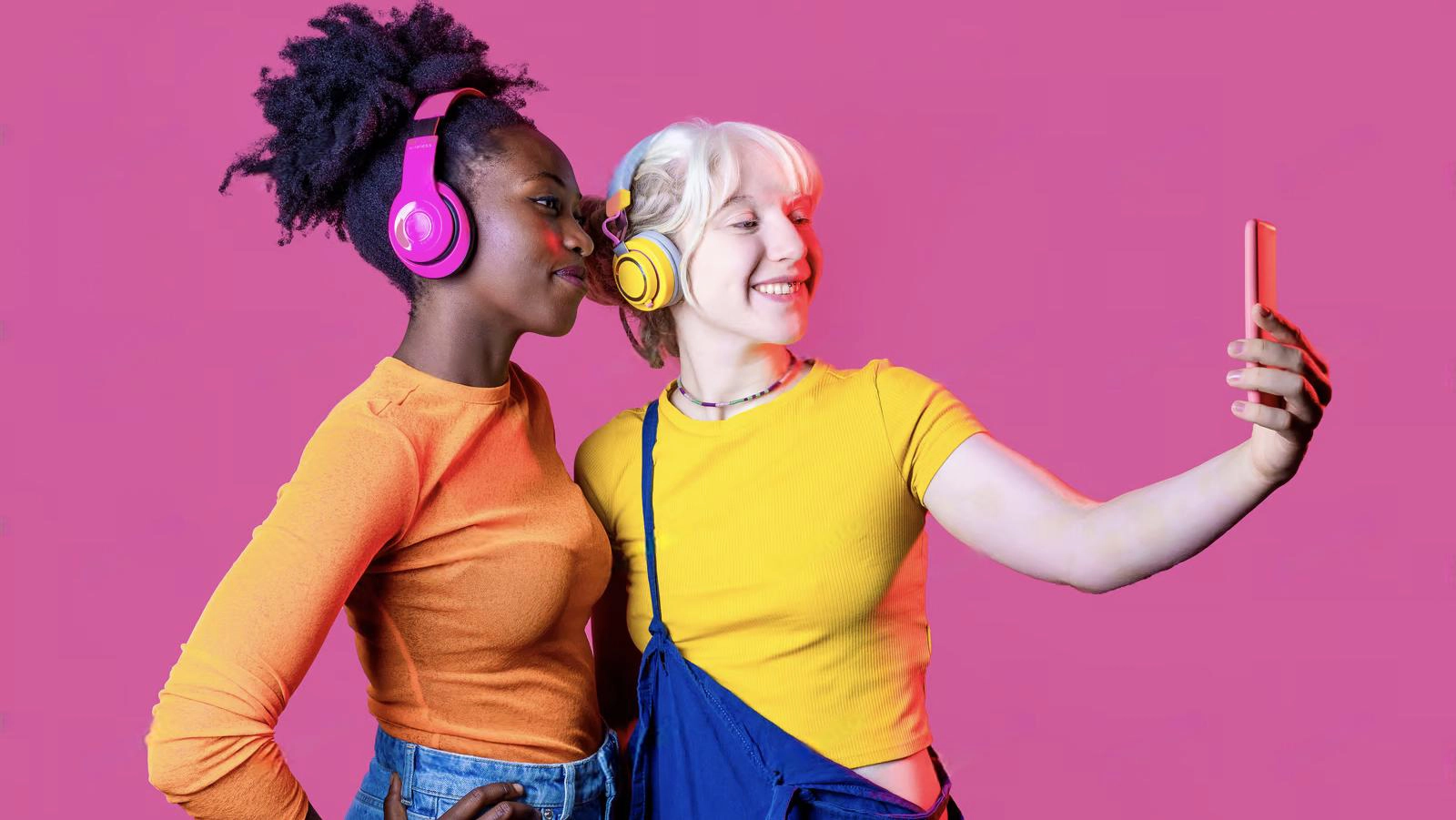 Two young women wearing headphones and colorful shirts take a selfie together against a solid pink background, capturing the perfect moment and showing off their understanding of fomo meaning in text.