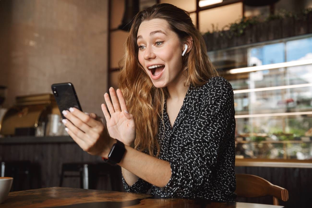 A woman sits in a cafe, smiling and waving at her phone during a video call. She wears wireless earbuds and a smartwatch—perfect accessories for anyone who loves tech reviews.