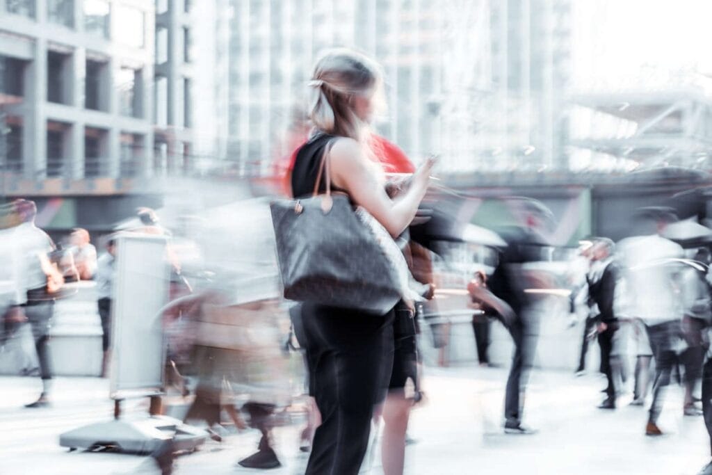 Blurred image of a woman with a large handbag standing among moving people in an urban area, subtly illustrating Marketing Social Proof amid modern buildings in the background.