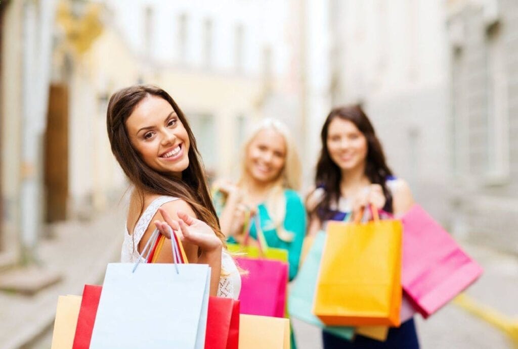 Three women stand outdoors holding colorful shopping bags, with one woman in the foreground smiling towards the camera—offering perfect social proof for effective marketing campaigns.