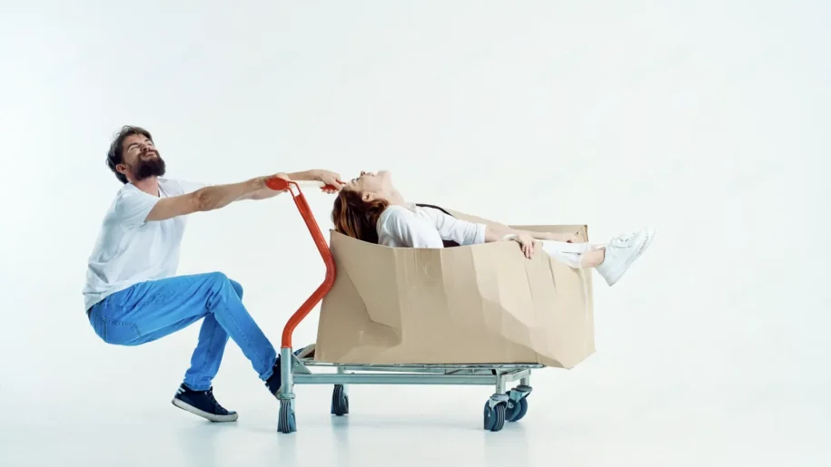 A man pulls a shopping cart with a woman lying inside a large cardboard box; their playful antics in a minimalist studio setting hint at creative ideas on how to reduce cart abandonment.