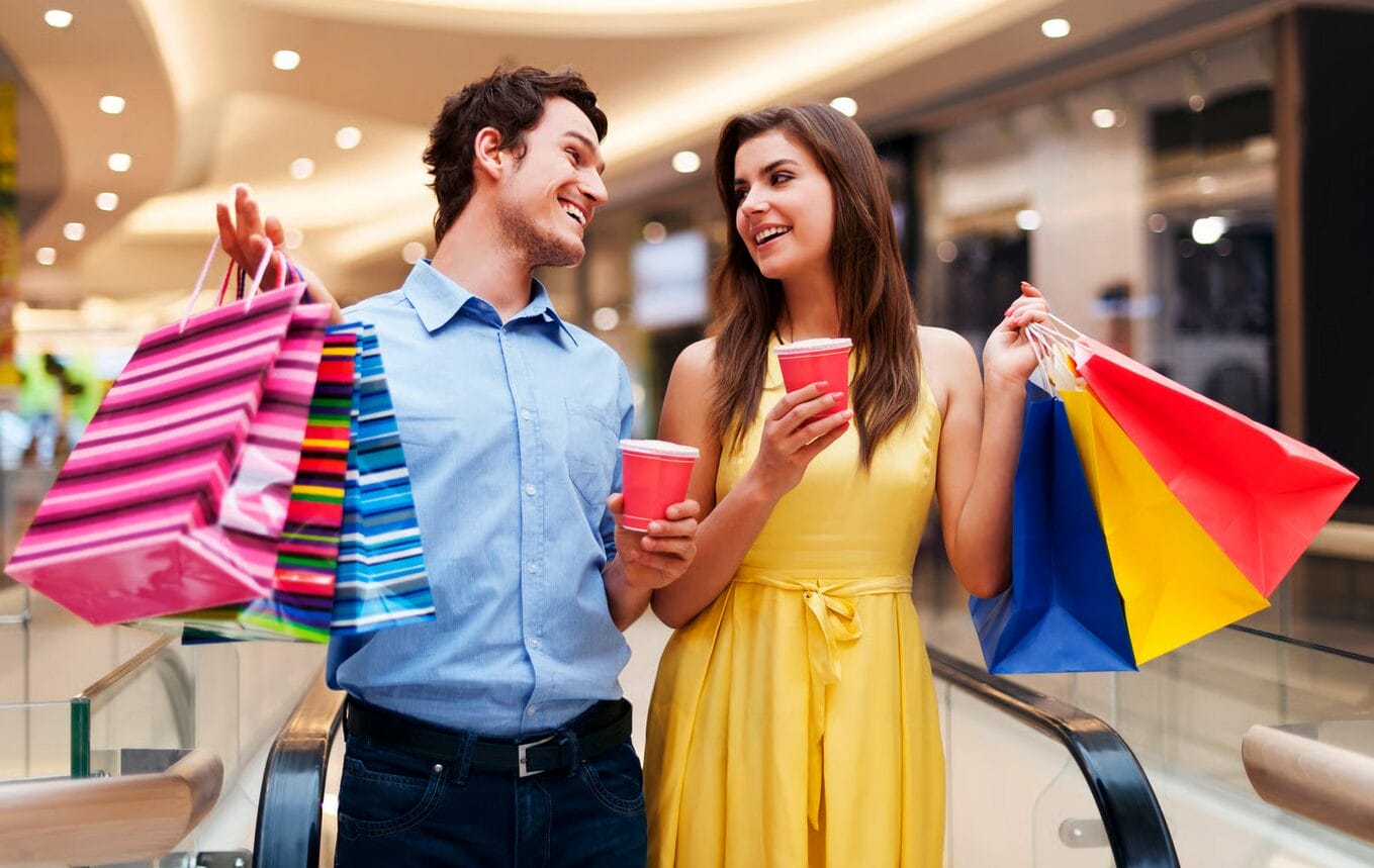 A man and a woman holding shopping bags and drinks stand together indoors, smiling at each other in a brightly lit shopping mall.