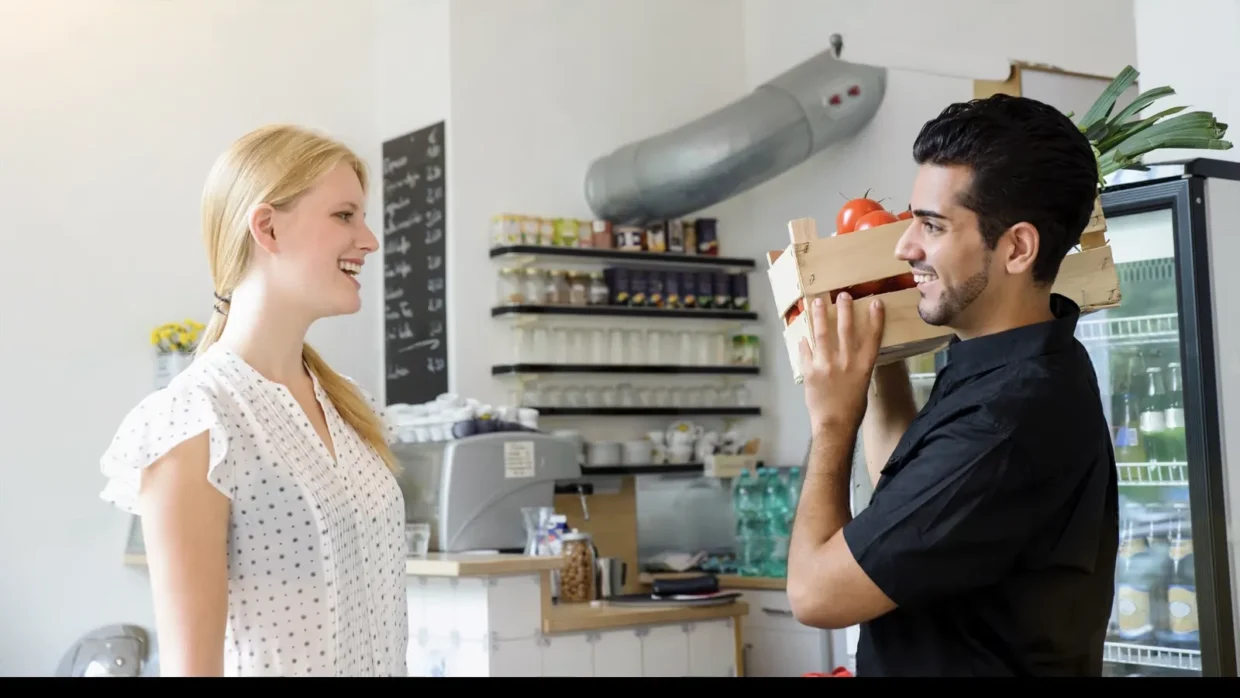 A man holding a crate of vegetables stands in front of a woman in a shop or cafe setting, where shelves with jars, bottles, and a refrigerator are visible—an ideal scene for applying customer feedback analysis methods.