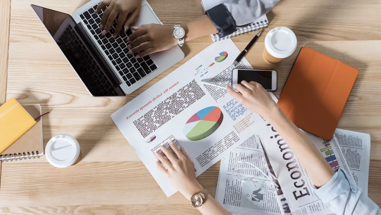 Two people work at a table with a laptop, charts, a notebook, coffee cups, a smartphone, and newspapers, discussing data and taking notes.
