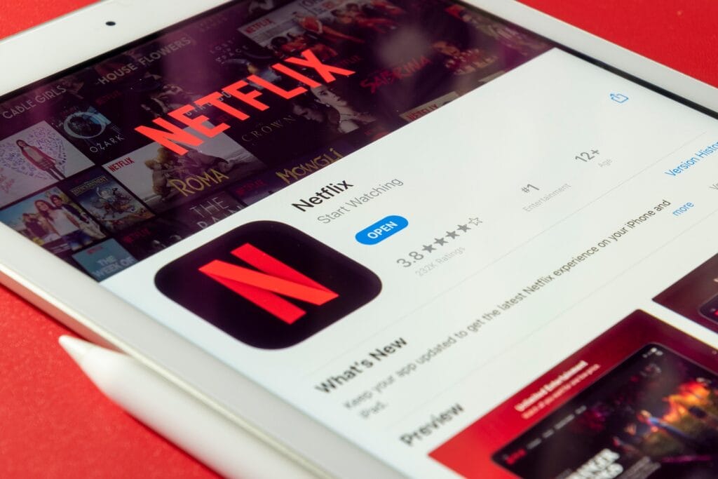 A tablet screen displays the Netflix app landing page in the App Store, showing the app icon, rating, and an "Open" button, with a background of Netflix show thumbnails.