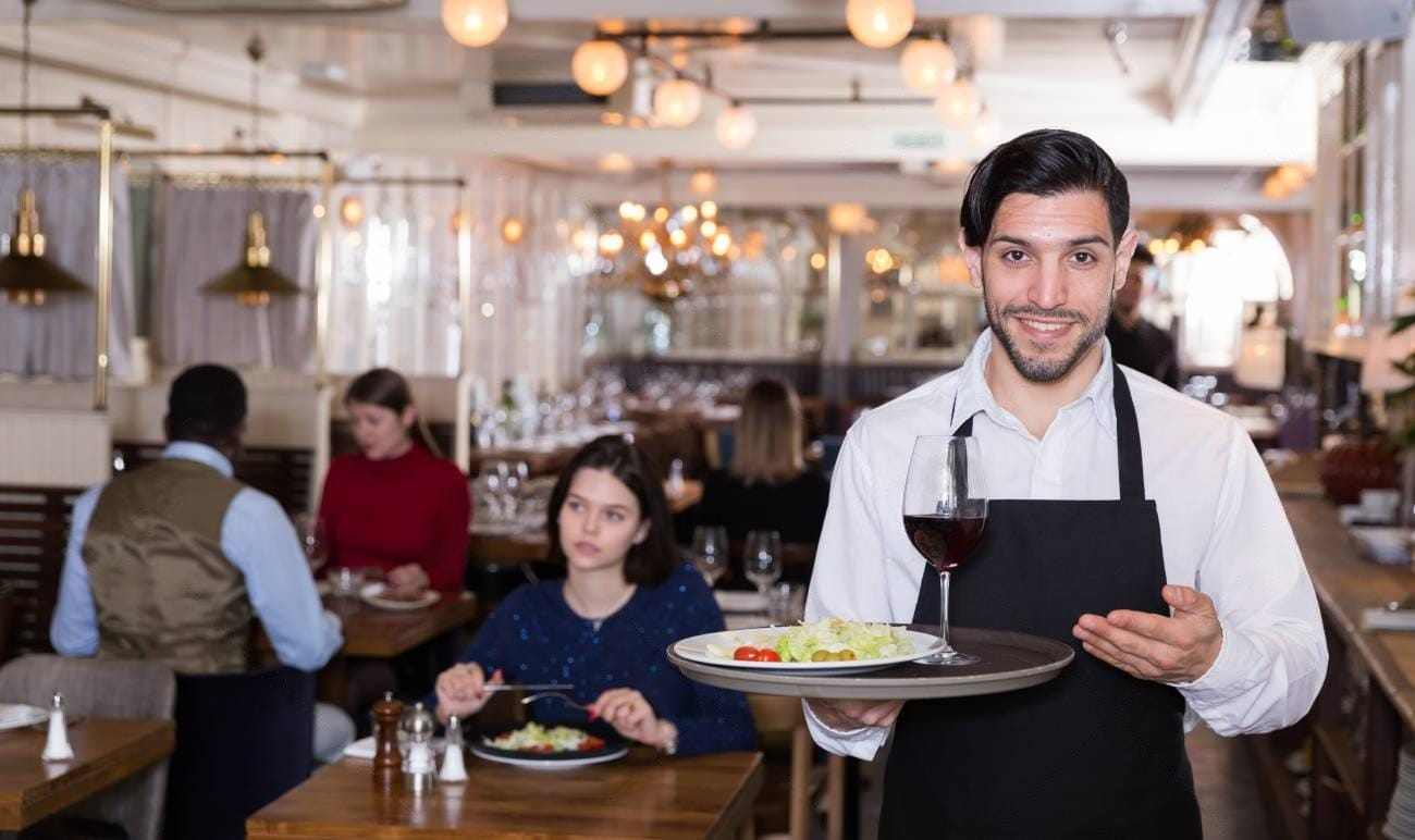 A waiter in a white shirt and black apron serves a plate of food and a glass of red wine in a busy restaurant with diners—capturing the perfect scene for restaurant review examples.