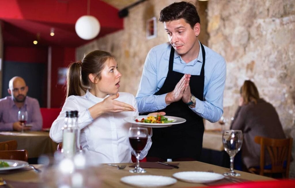 A woman seated at a restaurant points at her food and appears upset while a waiter listens and gestures apologetically, creating a scene often featured in Restaurant Review Examples.
