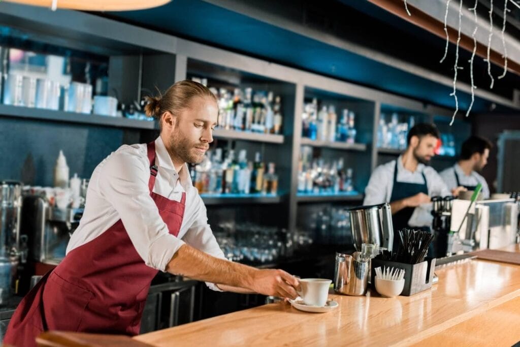 Bartender in a red apron places a cup of coffee on a wooden bar counter, creating the perfect scene for restaurant review examples, while another bartender works in the background of a modern bar setting.