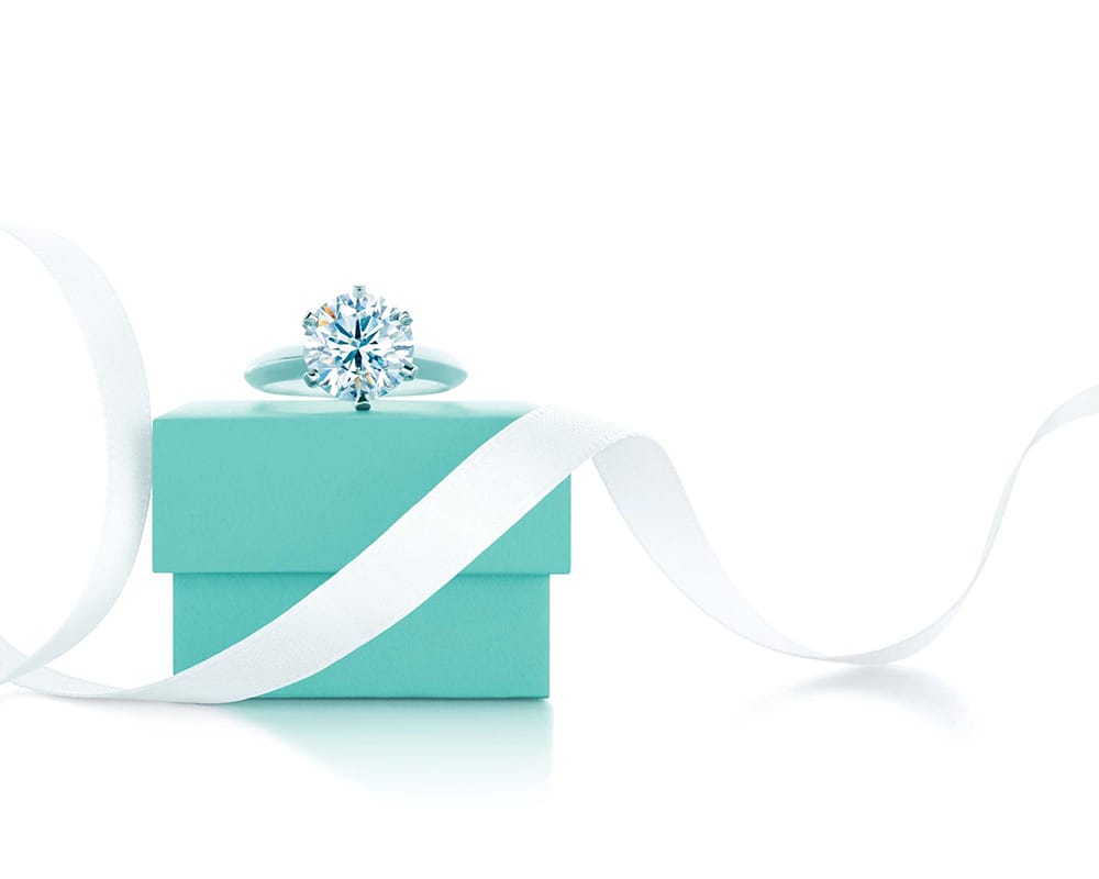 A diamond solitaire ring sits on a small turquoise box, perfect for a splash page or landing page, with a white ribbon curling beside it on a white background.